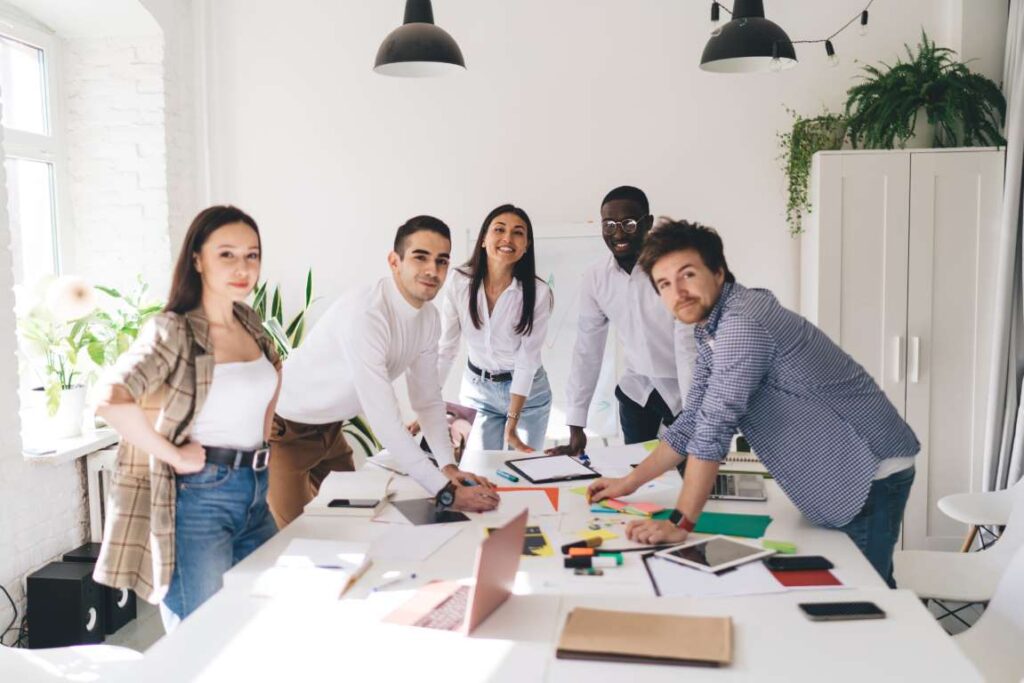 Group of multiethnic businesspeople in casual wear standing near table with gadgets and project documents and looking at camera in light creative office
